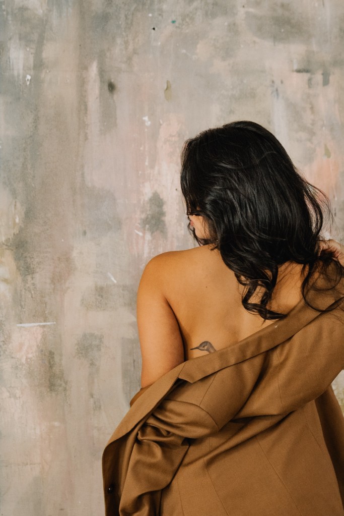 Woman with long black hair with her back to the camera wearing an off the shoulder brown jacket